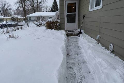 A shovel-width path across a snow-covered front yard to a side entry door, snow walls on either side and a shovel leaning against the siding