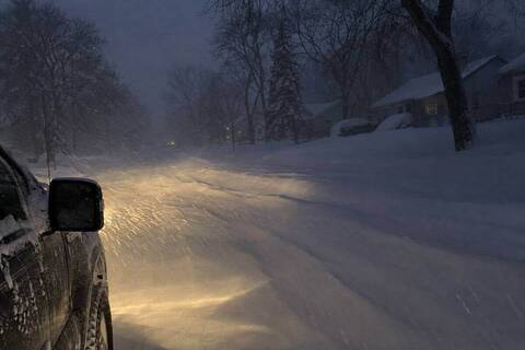 Truck headlights on an unplowed residential street during blowing snow before dawn, snow streaking through the beams