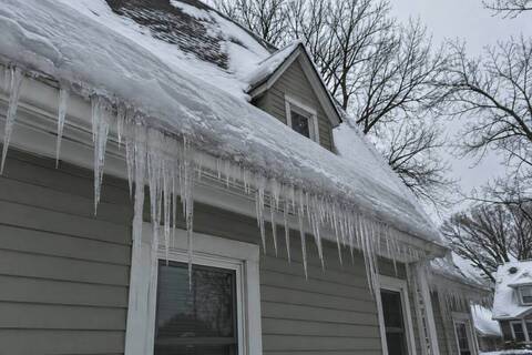 Thick icicles hanging from the gutter edge at the eave of a 1.5-story house, a ridge of ice built up along the roof edge above them, bare branches behind
