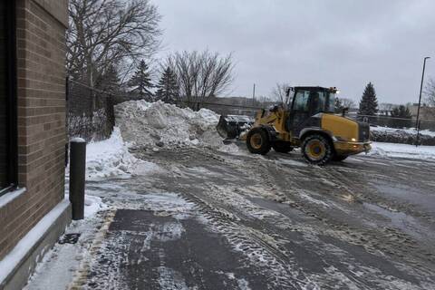 A compact wheel loader with a plain bucket at a snow pile at the back of a commercial parking lot, gray-white snow banked against a chain-link fence