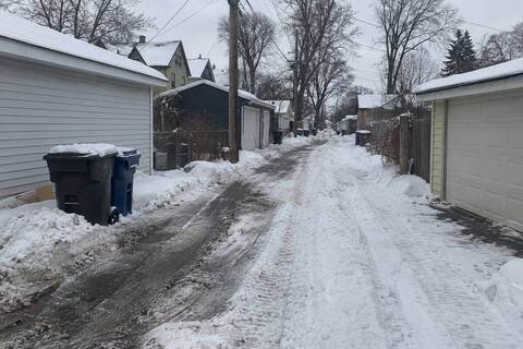 A narrow residential alley between detached garages, one track down to the surface and deep snow to the side, garbage carts pushed back against a garage wall