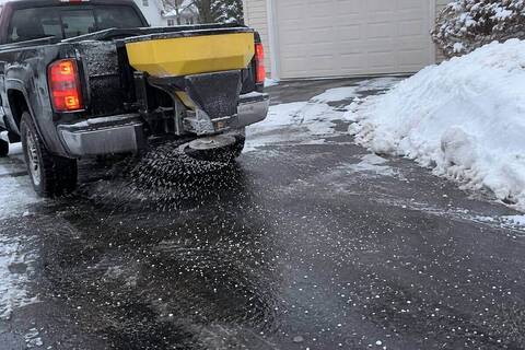 A tailgate spreader on the back of a pickup throwing granular ice melt across a wet driveway, pellets scattered across the dark pavement