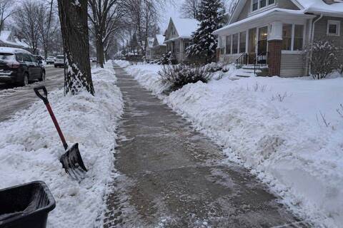 A public sidewalk past a city bungalow down to bare concrete across its full width, a shovel standing in the boulevard snowbank, snow piled on both sides