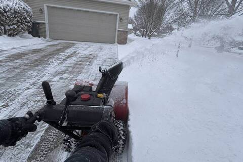 A walk-behind snowblower on a driveway with its chute turned to the side, a cut line in the snow behind it, gloved hands on the handles
