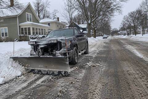 A pickup with a straight plow blade standing at a residential curb, blade resting on the pavement, snow-covered street and bare boulevard trees behind