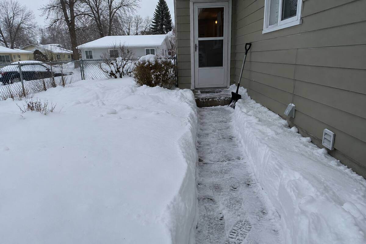 A shovel-width path across a snow-covered front yard to a side entry door, snow walls on either side and a shovel leaning against the siding