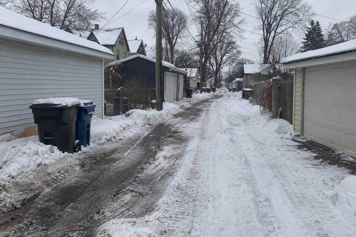 A narrow residential alley between detached garages, one track down to the surface and deep snow to the side, garbage carts pushed back against a garage wall