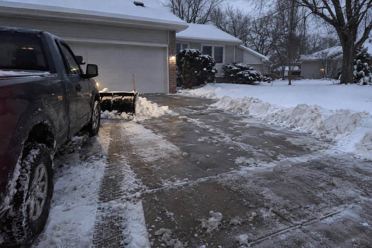 A pickup plow blade partway along a concrete driveway, snow curled off the blade edge and tire tracks behind it, closed garage door beyond