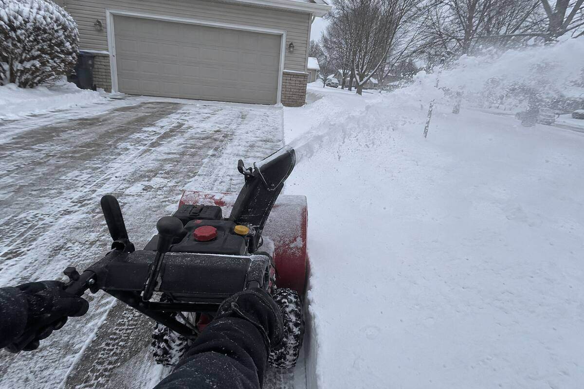 A walk-behind snowblower on a driveway with its chute turned to the side, a cut line in the snow behind it, gloved hands on the handles
