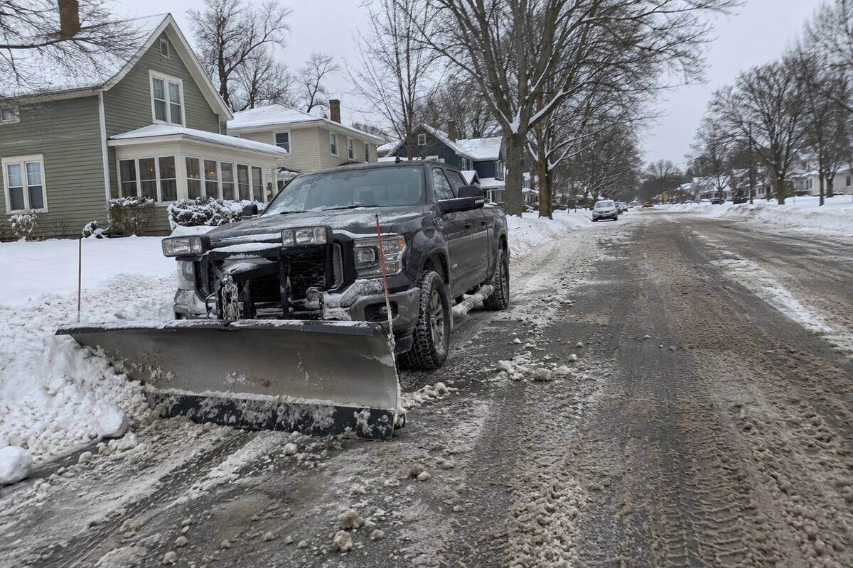 A pickup with a straight plow blade standing at a residential curb, blade resting on the pavement, snow-covered street and bare boulevard trees behind